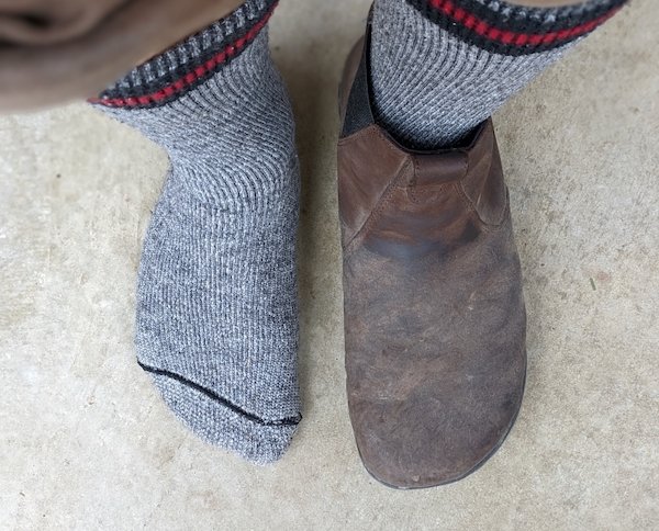 a top down view of a pair of feet standing on concrete. One is wearing a Lems Chelsea boot the other is in a sock to show that the shoe matches the shape of the bare foot