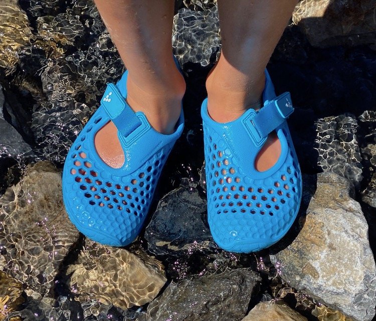 A close up of a child's feet standing on rocks in a stream of water wearing blue Vivobarefoot Ultra barefoot sandals water shoes