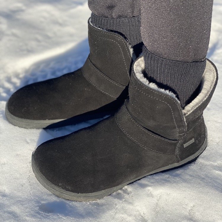 A close up side view of a pair of feet wearing black Be Lenka Polaris sheepskin warm barefoot winter boots in snow