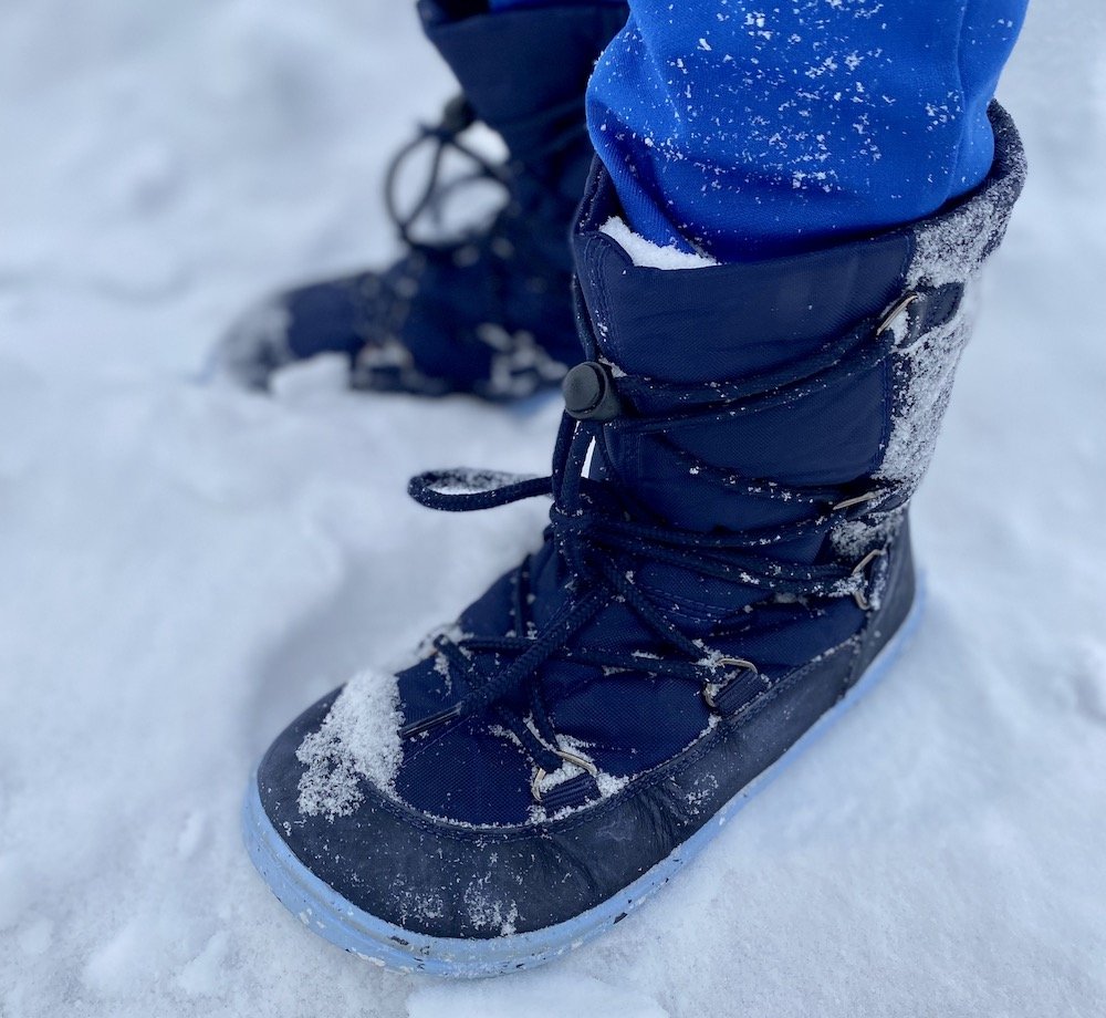 A close up of a child standing in snow wearing Be Lenka Snowfix kids barefoot winter boots