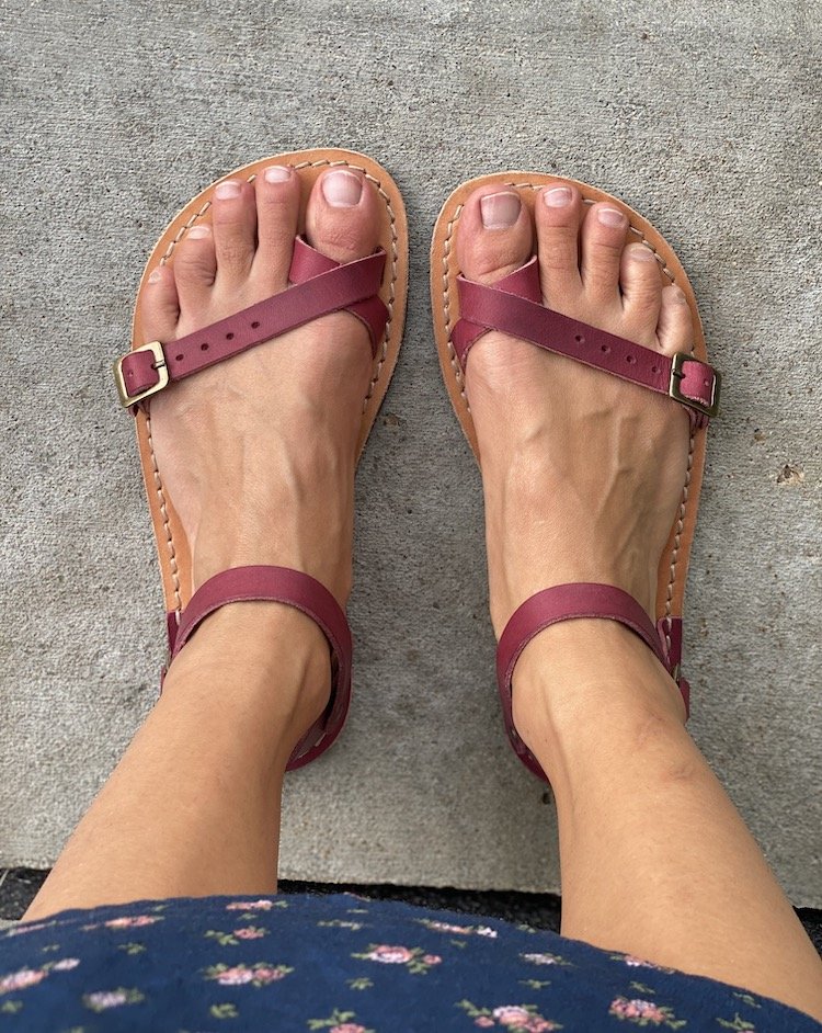 A top down view of a pair of feet standing on concrete wearing pink Crupon Nomade barefoot sandals with the bottom hem of a floral dress visible