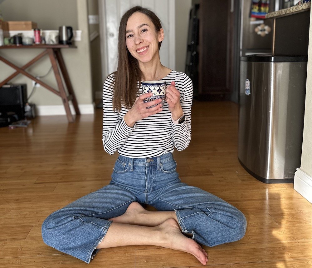 A woman sitting on the floor cross legged holding a cup of tea smiling at the camera