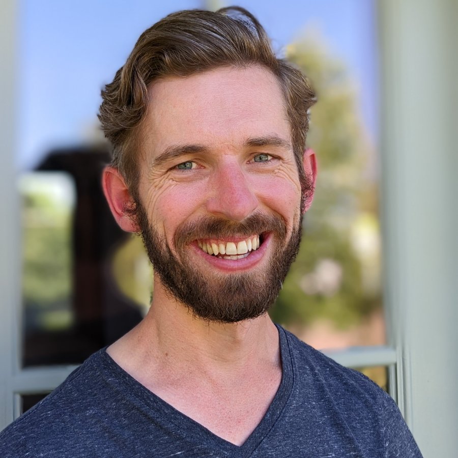 A white man smiling at the camera with blue eyes, a blue v neck shirt, and wavy hair