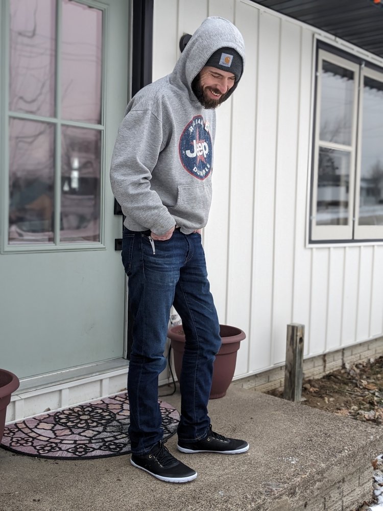 Full body photo of a white bearded man wearing a hoodie and jeans looking down at his Tolos Athletic barefoot sneakers in black with a white zero drop abrasion resistant thin outsole