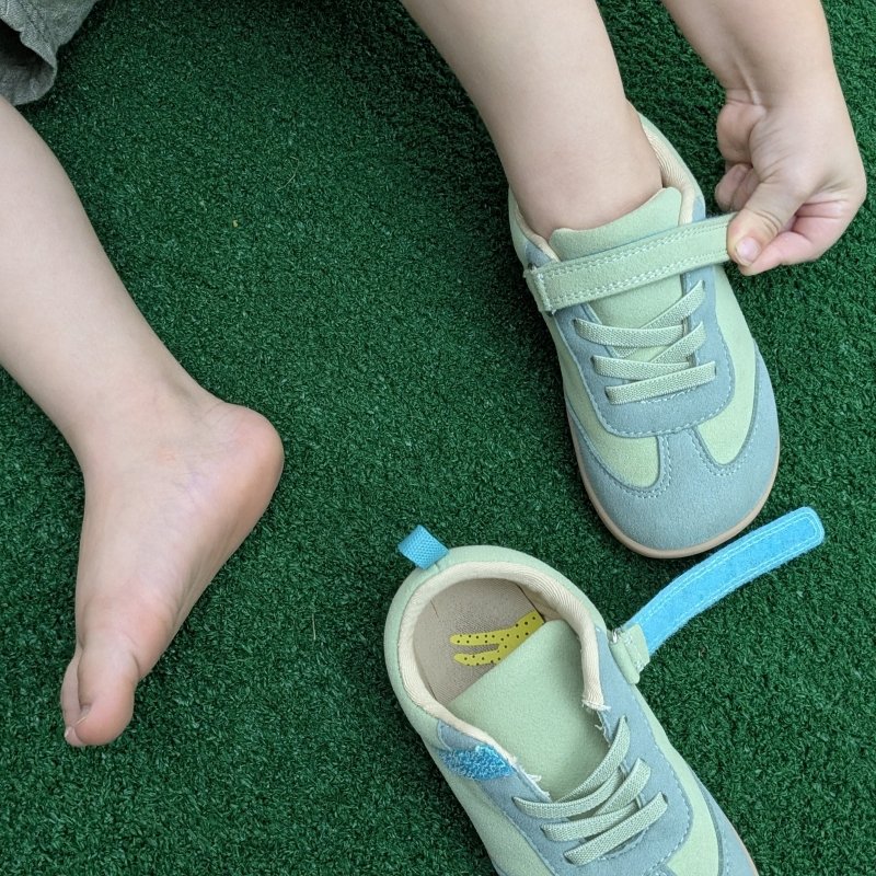 Top down view of a young toddler putting on a pair of green retro vegan suede wide toe box sneakers from Ten Little. One foot is bare, and his little hand is closing the velcro on the other shoe