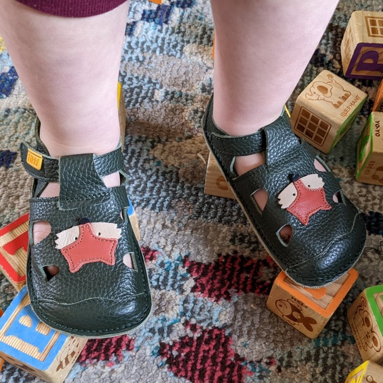 Tikki Nido sandals being worn by a toddler standing on wooden blocks They have a very flexible rubber sole for new walkers