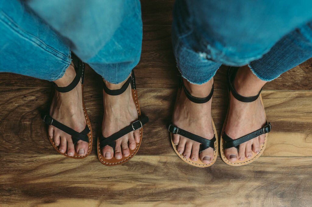 two pairs of feet standing side by side both wearing black Crupon sandals Nomade, with one in barefoot width and the other in Extra wide width