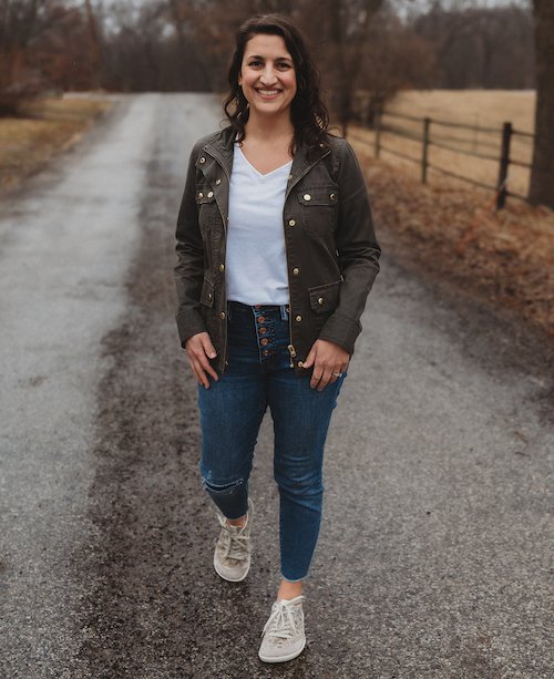 A woman with wavy brown hair wearing jeans, a white tshirt, and olive green field jacket walking towards the camera smiling. On her feet are comfortable extra wide Groundies Amsterdam sneakers with gold floral pattern