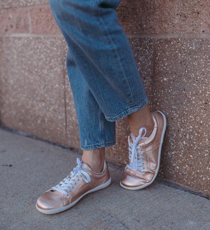 Close up of a pair of feet wearing Rose Gold Groundies metallic sneakers with white laces. One foot is propped up on the wall and the toes are flexed showing the sole flexibility. The model is wearing mom jeans