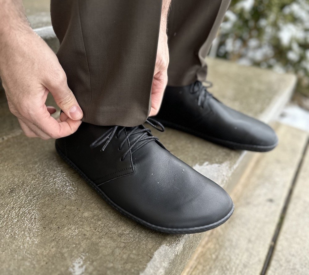 A side view of a man wearing dress pants and black groundies Milano barefoot dress shoes desert boots in wide Barefoot+