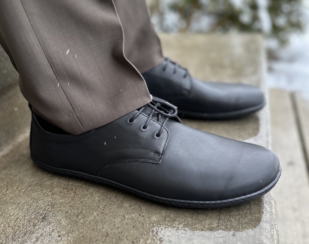 a man standing on concrete wearing dress pants and black Groundies Palermo barefoot dress shoes.