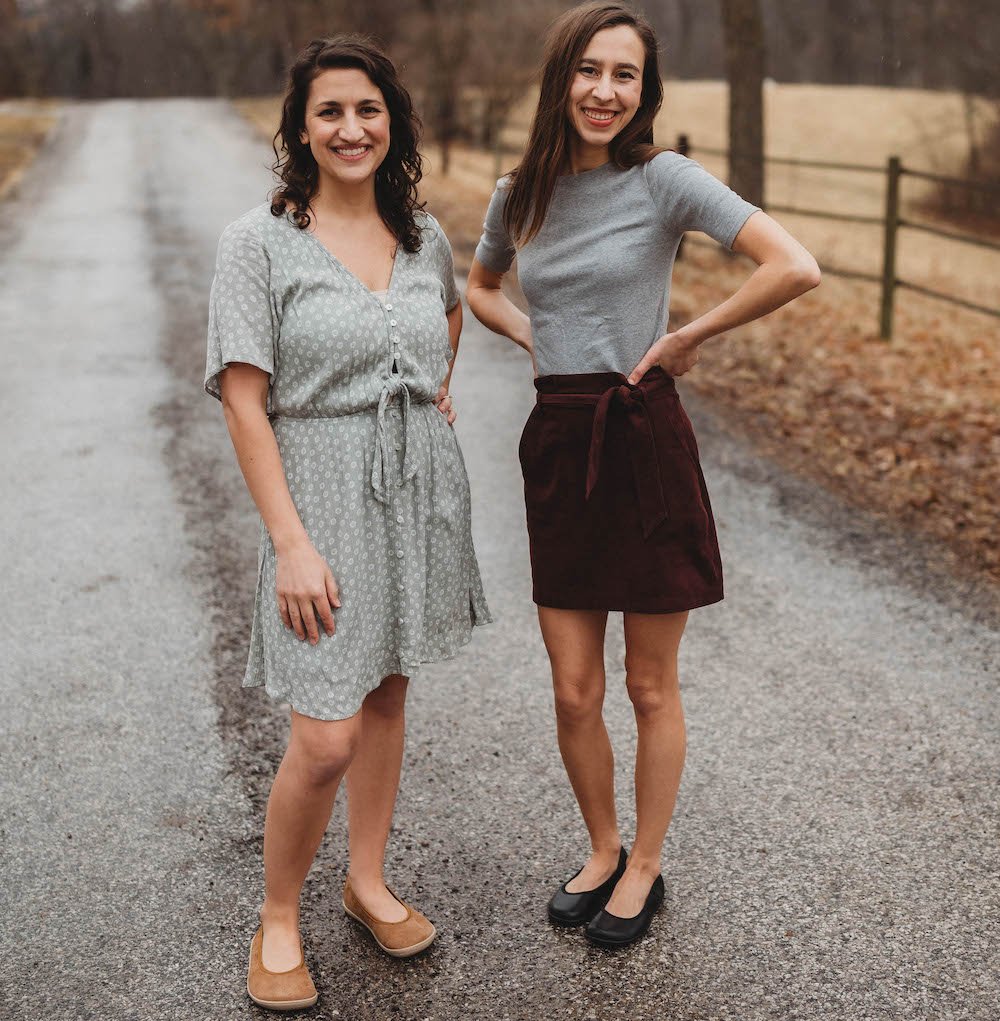 Two women standing in a road wearing dress clothes smiling at the camera. Groundies Lily ballet flats in two widths for wide and extra wide feet with anatomical toe box and zero drop outsole