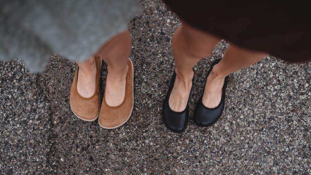 Top down view of Groundies Lily barefoot ballet flats in regular barefoot width on the right, and Barefoot Plus width on the left. Women's dress clothes are blurred in the foreground, and the focus is on the stylish ballet flats