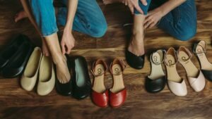 2 women sitting on the floor in jeans trying on various barefoot flats including Groundies Lily and Shapen Poppy. The women have different foot types, but are each finding the perfect fit from Anya's Shop