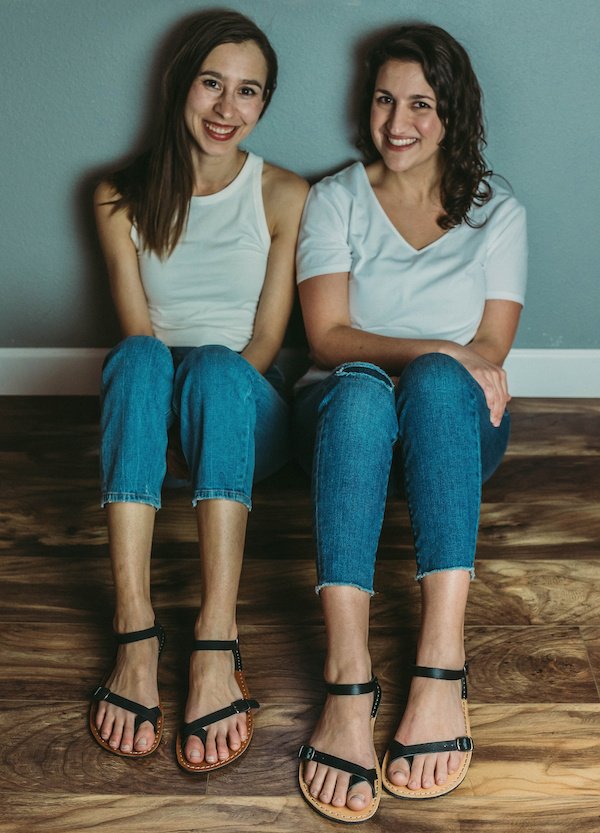 Two women sitting next to each other on the ground wearing white tees and jens and black Crupon Sandals barefoot sandals