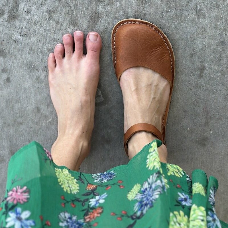 A close up of a pair of feet standing on concrete - the left foot is bare and the right foot is wearing Crupon Sandals Trevi barefoot in Ambra with a natural foot shape. the hem of a green floral skirt is visible.