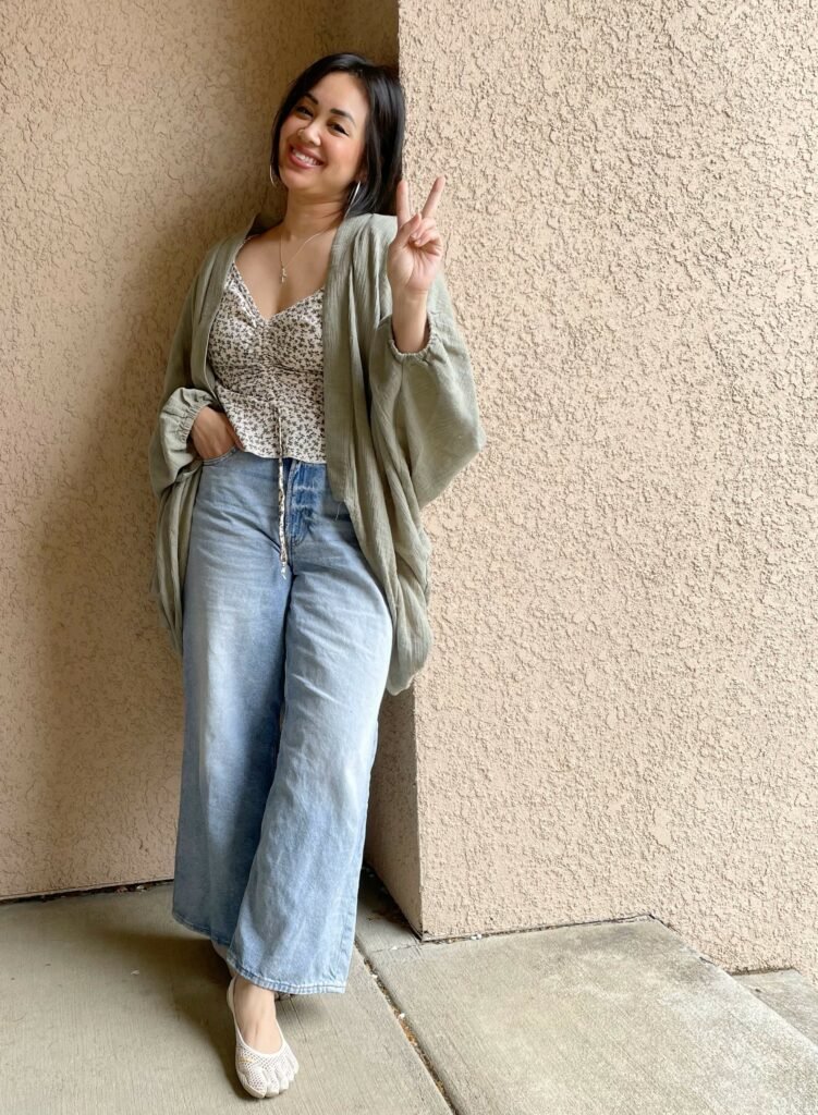 Heidi standing against a wall in wide leg light wash jeans, and airy cardigan kimono and vibram five fingers shoes