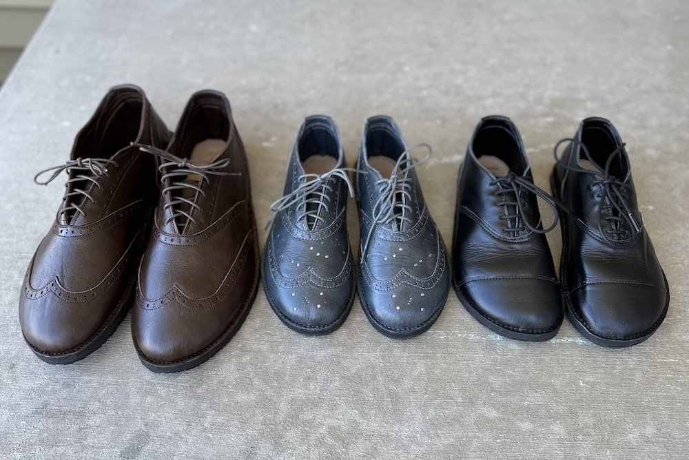 A row of Softstar Shoes Oxfords, barefoot dress shoes in black, brown, and grey, with wingtip brogue details