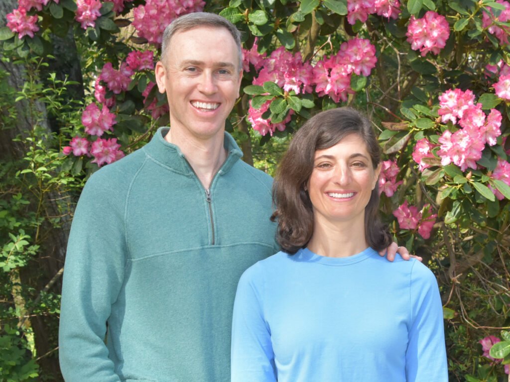 A man and woman from natural footgear standing together in front of a pink flowering bush. They are both smiling at the camera