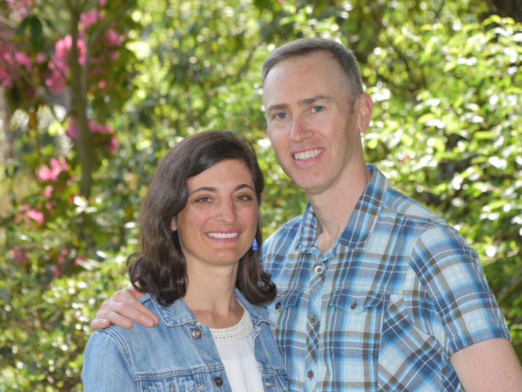 A brown haired woman Robyn and a gray haired man Marty from Natural Footgear standing with arms around each other in front of foliage. They are both smiling at the camera