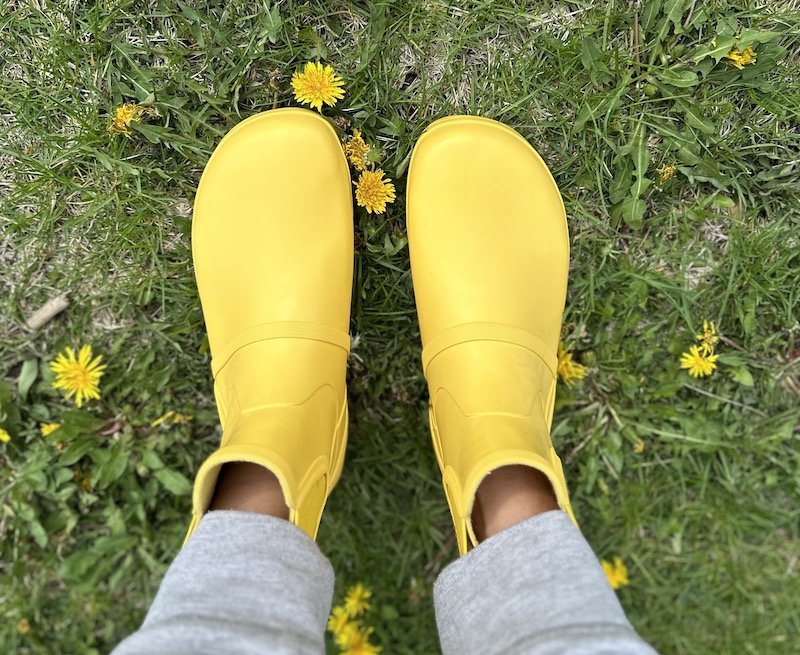 A top down view of a pair of feet wearing Xero Gracie rubber barefoot rain boots in bright yellow on grass