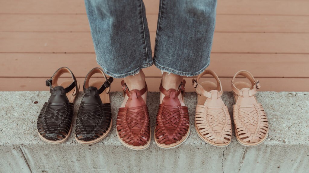 One person standing in a line up of 3 pairs of shoes, wearing the middle pair. The wide legs are showing off the Reddish Brown leather Sandal Huaraches by Anya. The black sandals are on the left, and the tan are on the right. All pairs have adjustable ankle straps and anatomical toe boxes