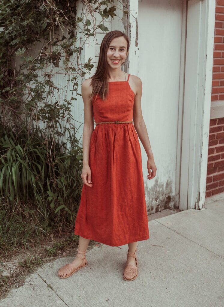 A woman in tan leather slips on with a wrapped up ankle strap wearing a burnt orange sun dress standing near a white shed with vines growing up the side. Her shoes are shaped like feet to allow natural foot functional while still looking stylish