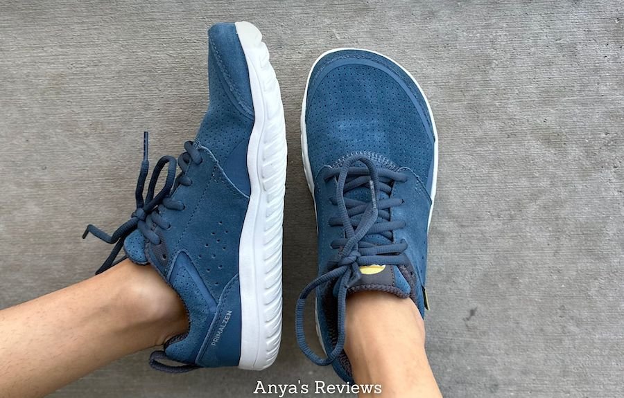 A close up view of a pair of feet resting on concrete wearing Lems Barefoot shoes Primal Zen suede in blue with the text "Anya's Reviews" on the bottom