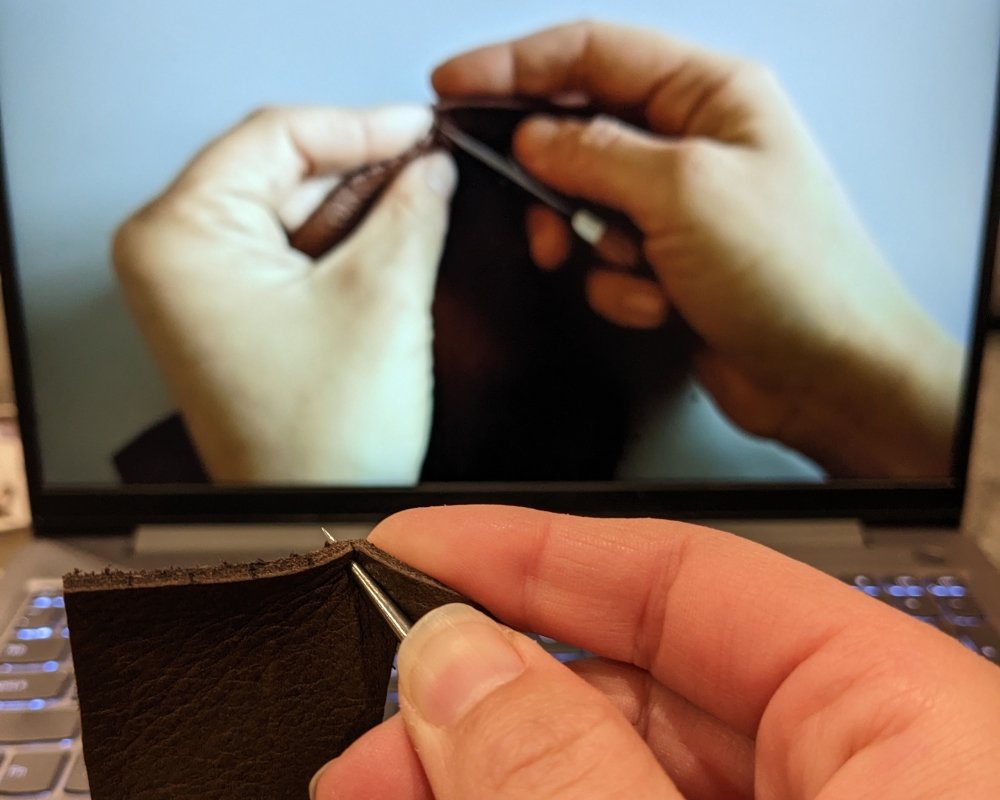 Close up of a hand holding an awl punching holes in a piece of leather with a computer screen playing an Earthing Moccasins instructional video in the background