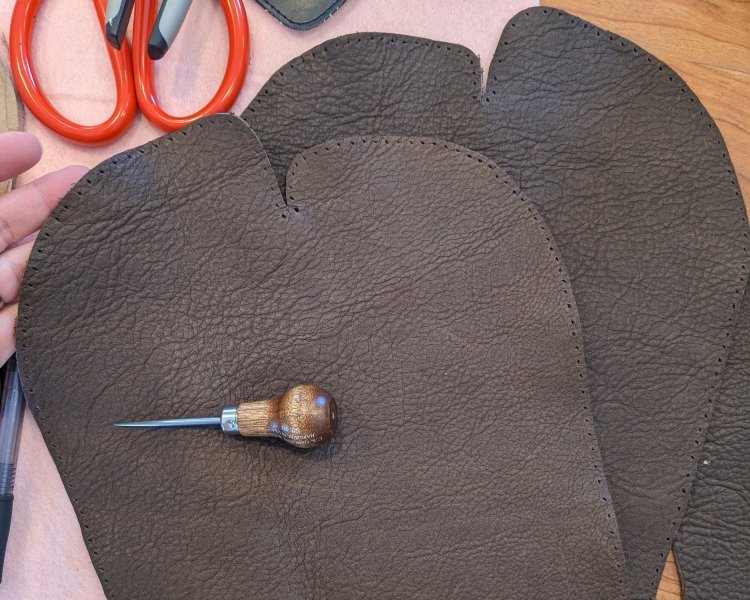 2 moccasin patterns cut out, a leather hole awl laying on top, and all 240 holes are visible around the outer edge