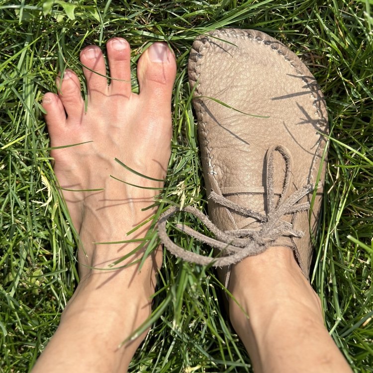 Top down view of someone's feet with 1 Earthing Moccasins Runner Mocc lace up full leather custom shoes standing on grass, and one bare foot to show the shape match