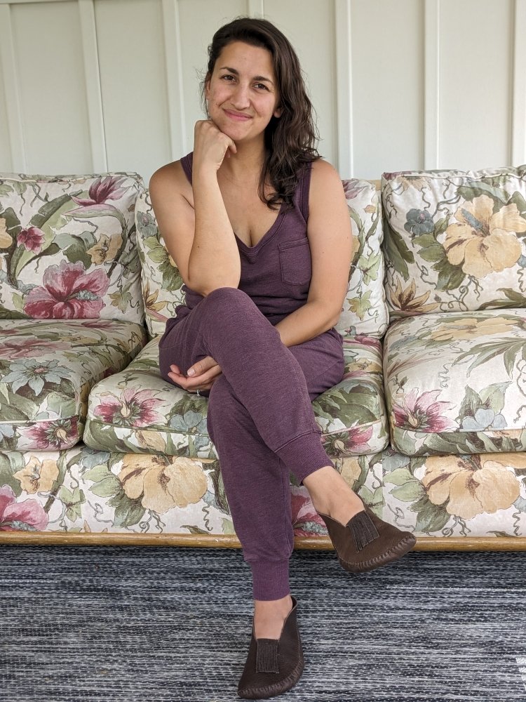 A woman with brown wavy hair in a purple jumpsuit sitting on a floral couch wearing brown leather handmade moccasins. She has her chin on her fist and is smiling slightly