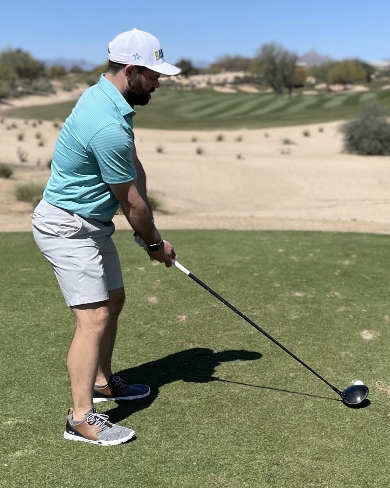 Full body photo of a man standing on the golf course about to take a swing. He is wearing shorts and polo, and zero drop comfortable minimalist golf shoes by TRUE Linkswear