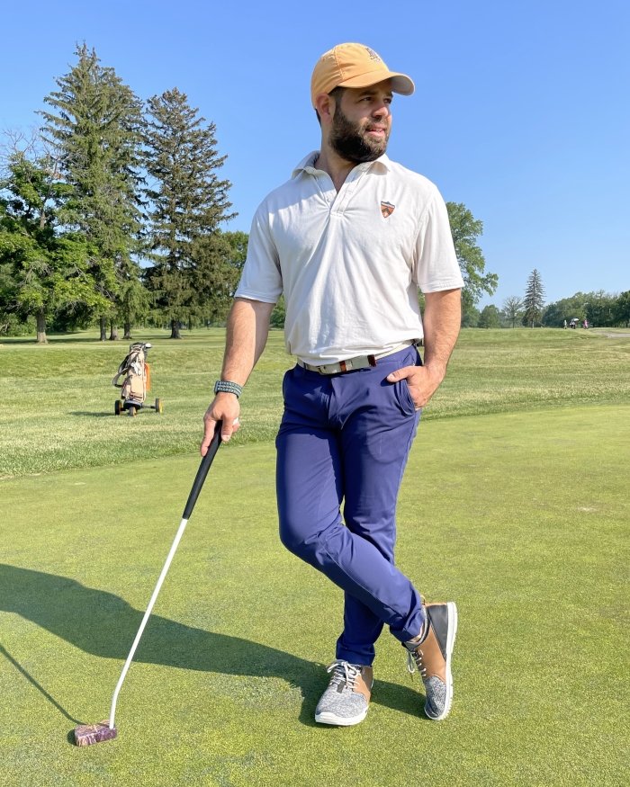 Full body photo of a man standing on the golf course wearing TRUE Linkswear OG Feel zero drop foot shaped golf shoes for proper alignment. He is leaning on his club with his hand in his pocket