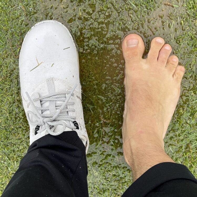 Top down close up of one bare foot and the other in True Linkswear Original 1.2 barefoot golf shoes for wet days with a wide toe box