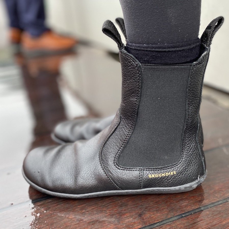 A close up of a pair of feet standing on a wet wooden deck wearing black Groundies Camden barefoot chelsea boots for autumn