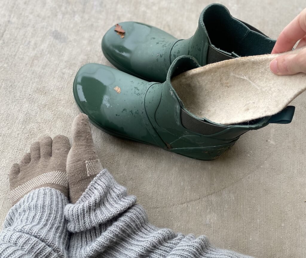 a pair of feet next to a pair of Xero shoes Gracie barefoot rain boots with a hand pulling a wool insole out of them