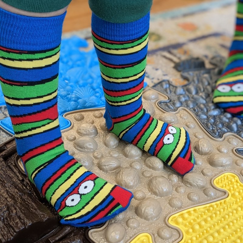 A young kid standing on a textured floor mat wearing Plus 12 striped anatomical socks that don't squish developing feet