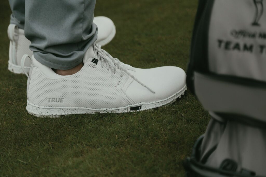 A side view of a person standing on a golf green wearing True Linkswear white Original 1.2 barefoot golf shoes with a golf bag next to his feet