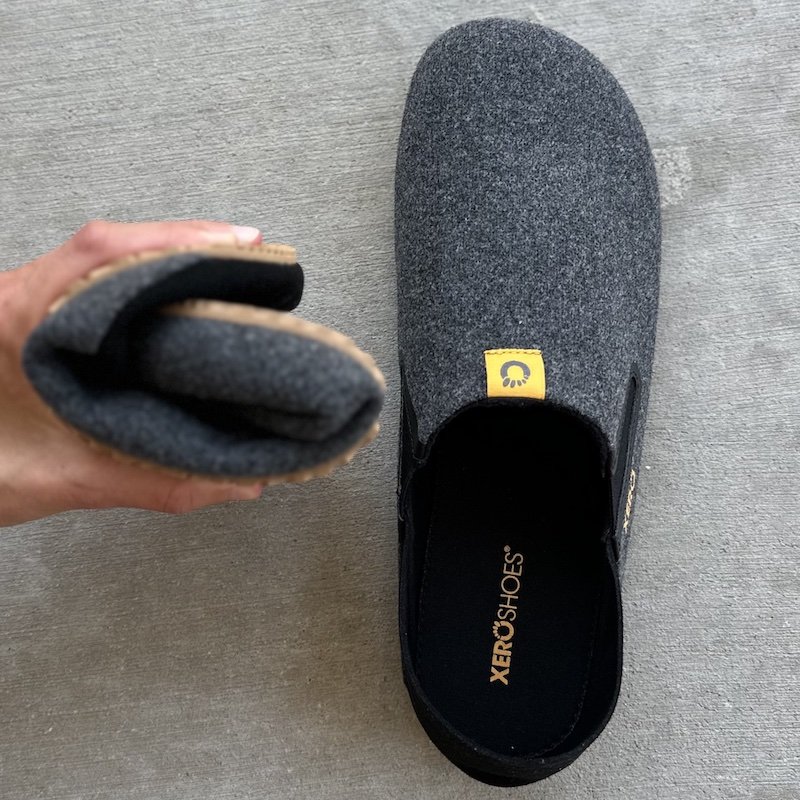 A top down view of a pair of Xero shoes barefoot slippers called the Pagosa in grey with a hand holding one shoe rolled up into a ball to show its flexibility