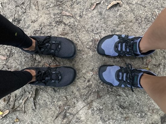 Top down view of two people standing toe to toe wearing Xero Shoes hiking shoes with wide toe boxes. Xero Scrambler Low and Mesa Trail WP with zero drop flexible outsoles are both being worn on a dirt path