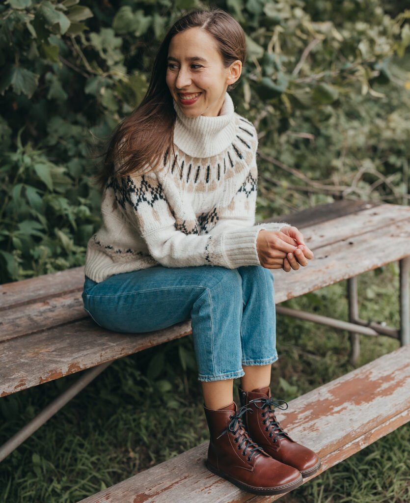 A woman sitting on a bench wearing a sweater and smiling wearing Peerko brown leather barefoot boots Go combat lace up style.