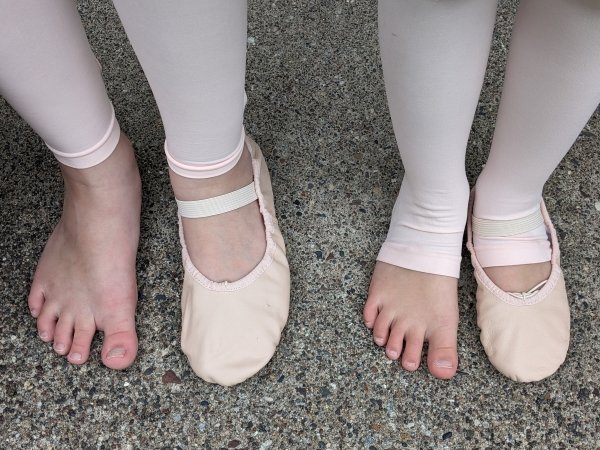 Top down view of two children's feet with 1 ballet shoe on, one shoe off. both wearing footless or stirrup tights to let their toes be free