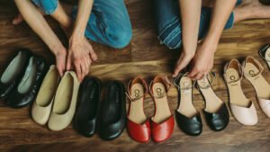 A row of pretty stylish women's barefoot dress shoes in a row on a wood floor with the arms of two people showing as they reach for the shoes