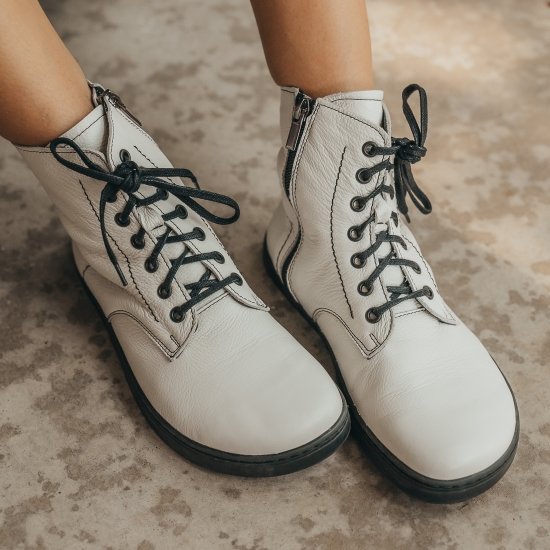 Close up image of a pair of legs wearing Peerko Go white leather combat style boots with black laces and a side zip. The wide toe box is prominent at the front of the image