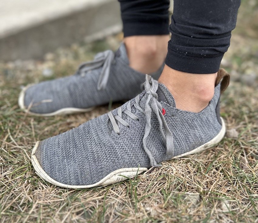 A close up side view of a pair of feet standing on grass wearing grey Wildling nebula barefoot shoes with a stretchy knitted wool upper