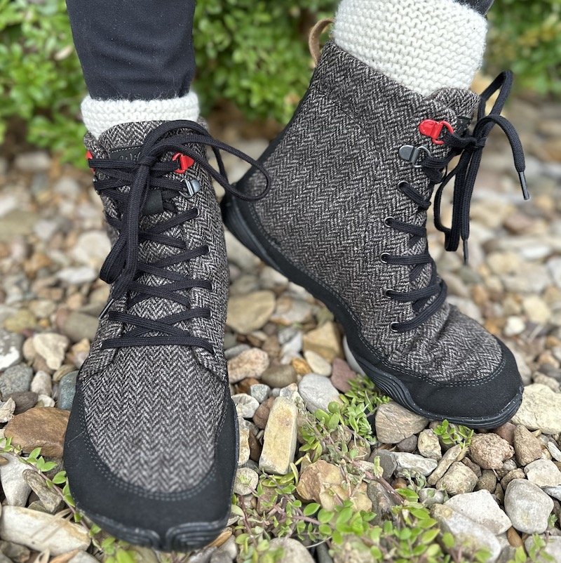 A close up of a pair of feet wearing Wildling Shoes North Wolf wool barefoot winter boots, standing on rocks with one foot turned out to the side.