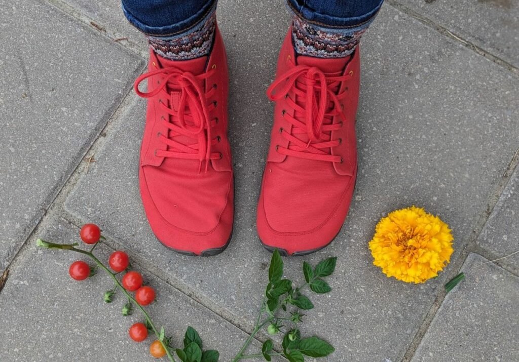 A top down view of a pair of feet wearing Wildling barefoot shoes Paprika standing on concrete with leaves and flowers surrounding them