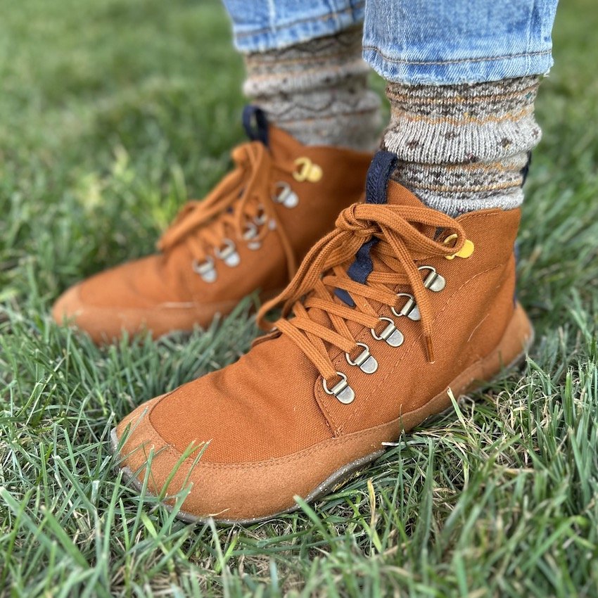 Front angled view of a person in caramel colored boots standing in the grass. Multicolored wool socks are visible above the shoe, which is Wildling Tejo Caramel with waterproof membrane and durable canvas uppers with quick lace eyelets and laces that match perfectly