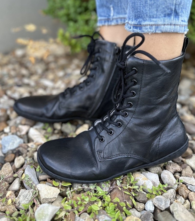 A close up side view of a pair of feet wearing the Mukishoes Igneous lace up barefoot combat boots in black leather with a wool lining.